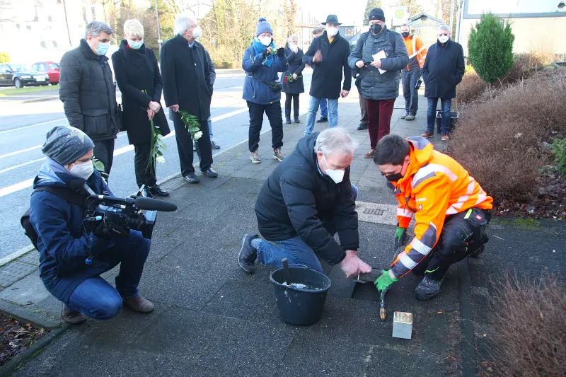 Eine Kamerafrau filmt zwei Mitarbeiter des städtischen Bauhofs bei der Verlegung des Stolpersteins, während die Bürgermeisterin und Historiker im Hintergrund zuschauen.