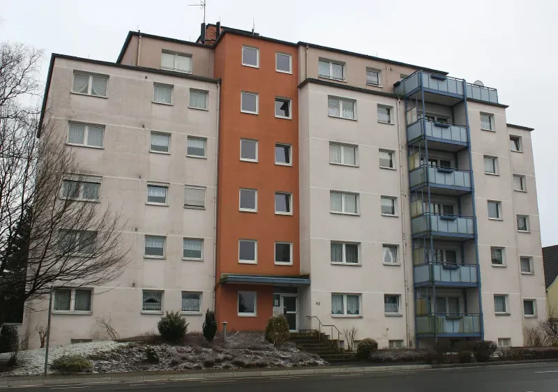 Ein sechsstöckiges, weiß verputztes Wohnhaus mit einem Flachdach. Der Bereich des Treppenhauses ist rot angestrichen; die Balkons sind blau.