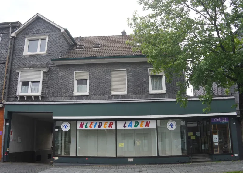 In einem zweieinhalbstöckigem verschiefertem Wohnhaus befindet sich im Erdgeschoss ein Ladengeschäft und links davon eine Durchfahrt in den HInterhof.