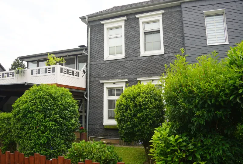 Vor einem zweistöckiges Wohnhaus mit einer Fassade aus Rheinischem Schiefer und einem großen Balkon befindet sich ein Vorgarten mit großen, grünen Sträuchern.