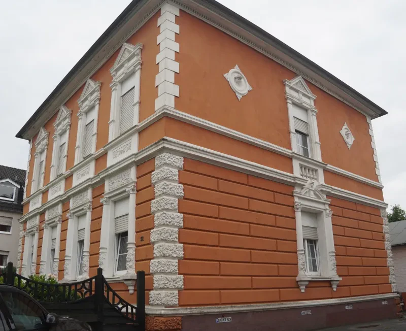 Ein zweistöckiges Wohnhaus im Jugendstil mit orangefarbenen Anstrich und weißen verzierten Fenstereinfassungen und Ecksteinen.