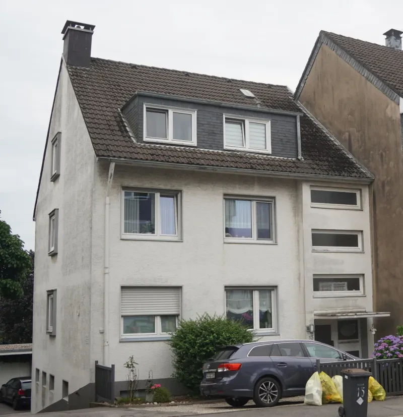 Auf einem nach hinten abschüssigen Hang steht ein weiß verputztes, dreistöckiges Wohnhaus mit dunkelgrauem Satteldach. In der oberen Etage befindet sich eine breite Dachgaube mit zwei Fenstern.