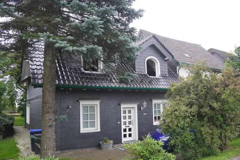 Ein kleines zweistöckiges Wohnhaus, dessen Fassade aus dunklem Schiefer besteht, ist von einem kleinen Garten umgeben.