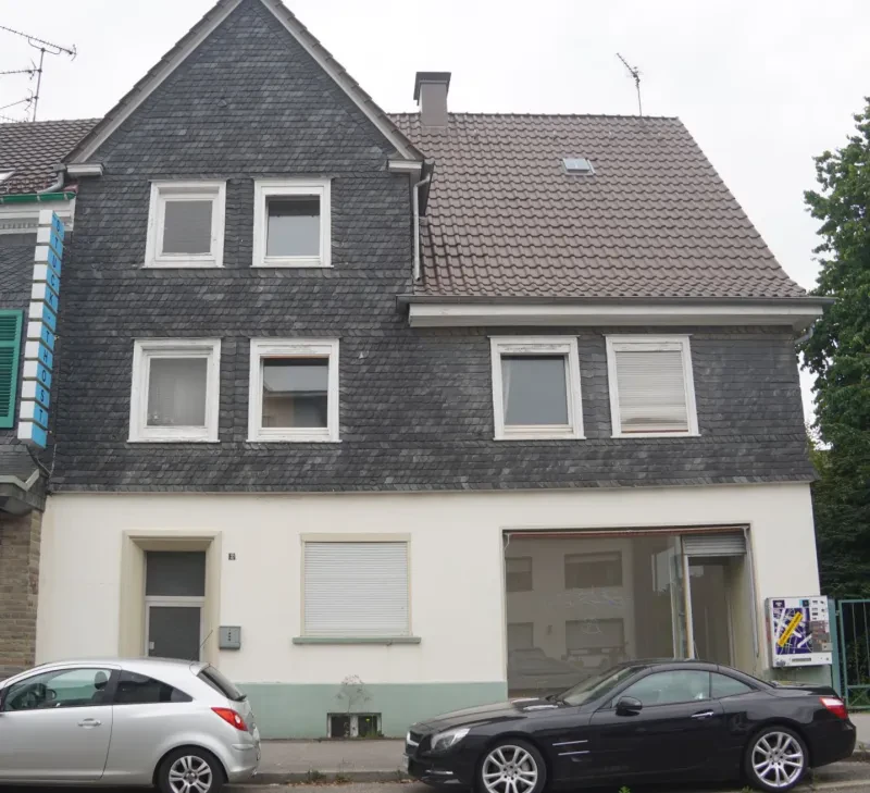 Ein dreistöckiges Wohnhaus mit Fassade und Dach aus dunklem Rheinischem Schiefer. Im Erdgeschoss befindet sich ein Ladengeschäft, dessen Wände weiß verputzt sind.