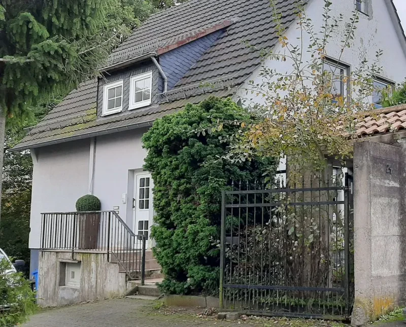 Ein geöffnetes Gitter gibt den Blick frei auf ein älteres, weißes Einfamilienhaus mit dunklen Dachziegeln