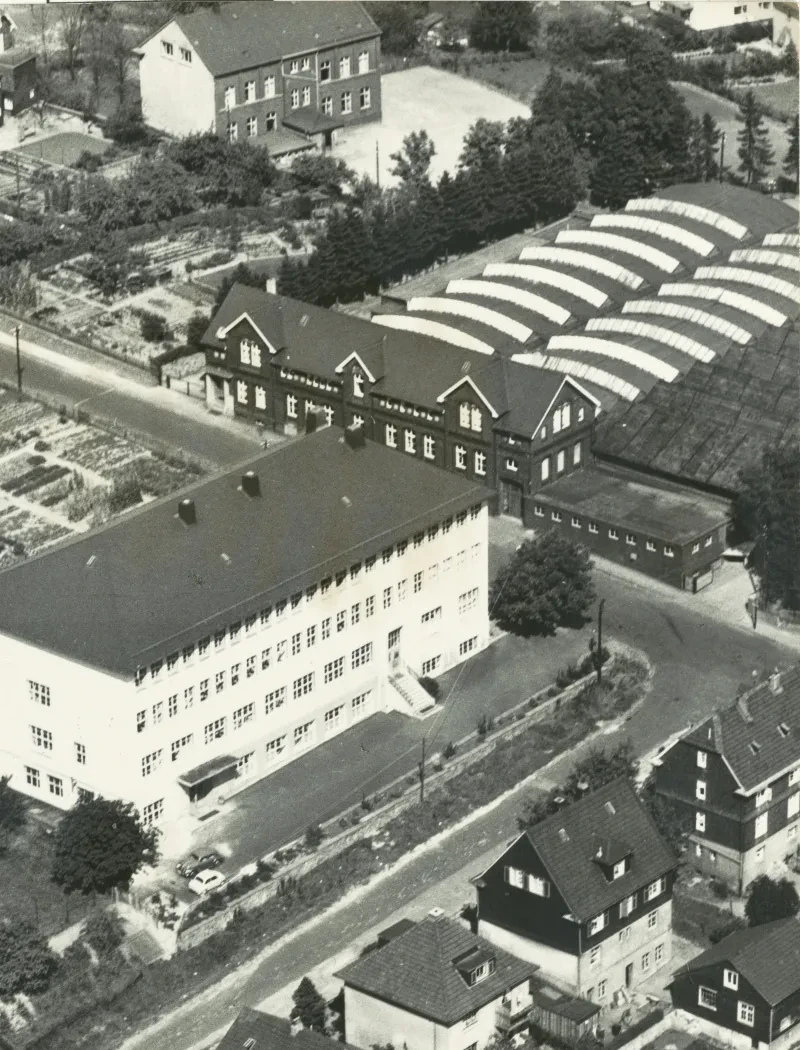 In einer Wohnstraße befindet sich auf der linken Seite eine dreistöckige Berufsschule und auf der rechten Seite ein zweistöckiges Fabrikgebäude mit daran anschließenden Produktionshallen.