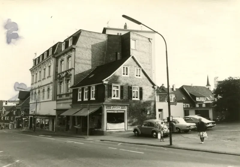 Ein kleines dreistöckiges Schieferhaus schmiegt sich in die Niesche von zwei dahinter stehenden vierstöckigen Gebäuden. Alle Gebäude haben zur Straßenseite Geschäfte mit Schaufenstern.