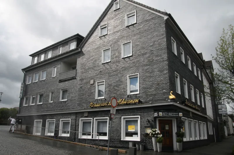 An einer Straßenkreuzung befindet sich ein großes, vierstöckiges Haus, dessen Fassade und Dach aus dunklem Bergischem Schiefer besteht. An zwei Seiten befindet sich die Inschrift "Restaurant zum Schwanen" und genau an der Ecke ist über der Eingangstür ein goldener Schwan zu sehen.