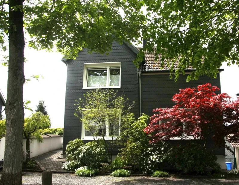 Ein zweistöckiges Wohnhaus hinter roten und grünen Sträuchern außen mit dunklem Bergischen Schiefer verkleidet.