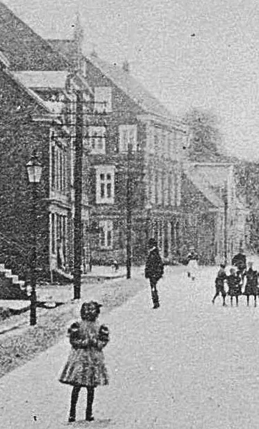 Vor einem vierstöckigen Schieferhaus stehen Straßenlaternen und Strommasten. Auf der Straße spielen Kinder.