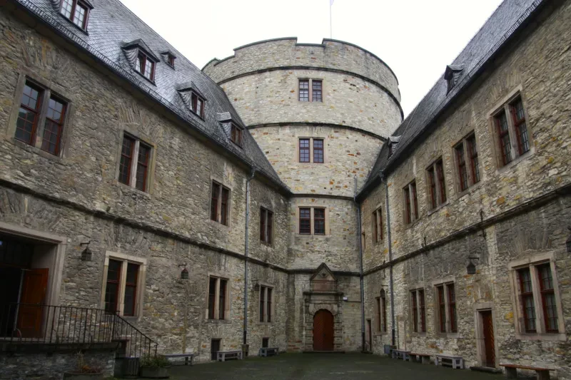 In einem Innenhof laufen zwei zweistöckige Gebäudeflügel aus grob gehauenen Steinen spitz auf einen breiten Turm mit Flachdach zu.