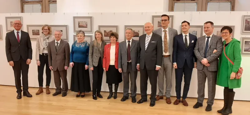 Sieben Männer und fünf Frauen stehen in einm Aussellungsraum. An der Wand hinter ihnen hängen 12 Aquarelle.