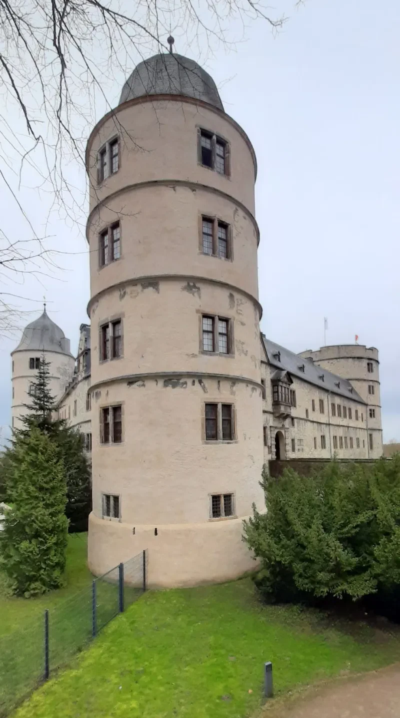 Ein vierstöckiges, helles Schloss mit Satteldach wird im Süden von zwei schmalen Türmen und im Norden von einem mächtigen Einzelturm flankiert.