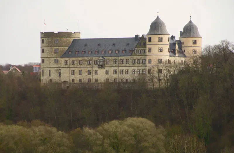 Von Südwesten blickt man über ein bewaldetes Gelände auf die helle Wewelsburg, die durch einen dreieckigen Grundriß und ein Satteldach auf den drei Flügeln auffällt.