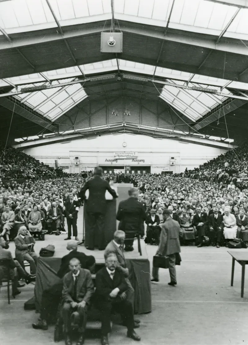 Eine Halle voller Menschen, im Vordergrund ein mit dem Rücken zur Kamera spricht ein Redner zum Publikum. Er steht auf einem Podest, davor sitzen zwei Männer.