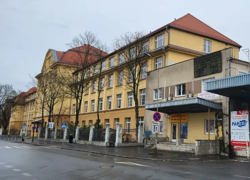 Rechts neben einem zweiflügeligen, vierstöckigen, ehemaligem Verwaltungsgebäude befindet sich eine Werkseinfahrt.