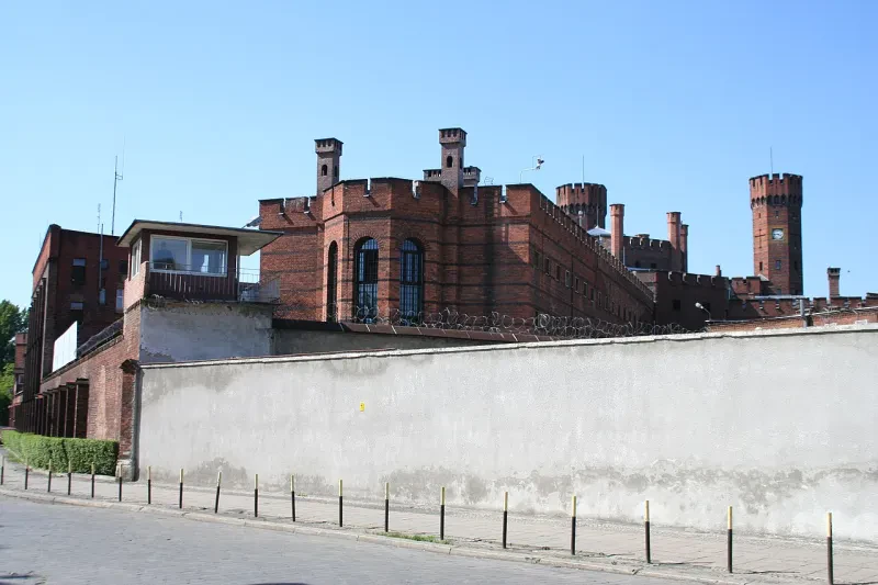 Hinter zwei Mauern und Stacheldraht ragt das rote Backsteingebäude eines alten Gefängnisses hervor. Die Eckmauer ist durch einen Wachturm gesichert.