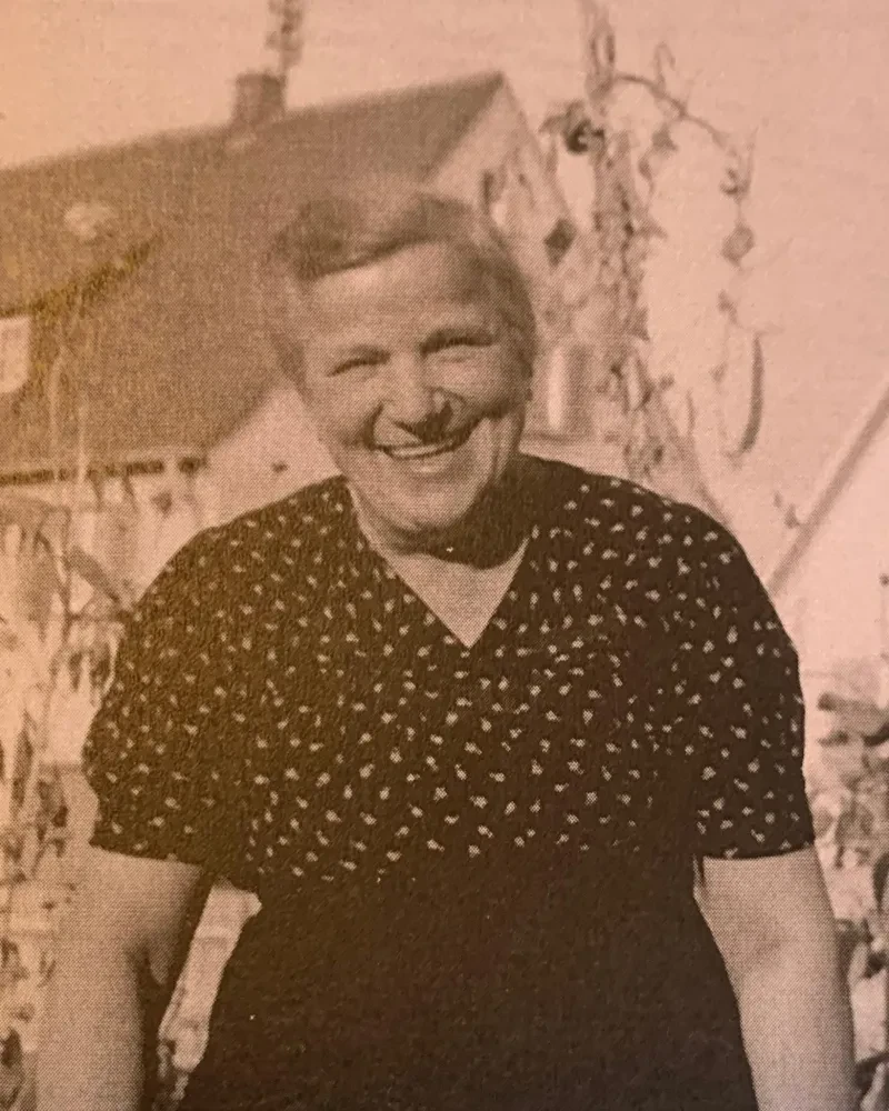Eine Frau in einer Bluse mit kurzen Ärmeln lacht in die Kamera. Sie arbeitet scheinbar im Garten. Im Hintergrund ein Haus.