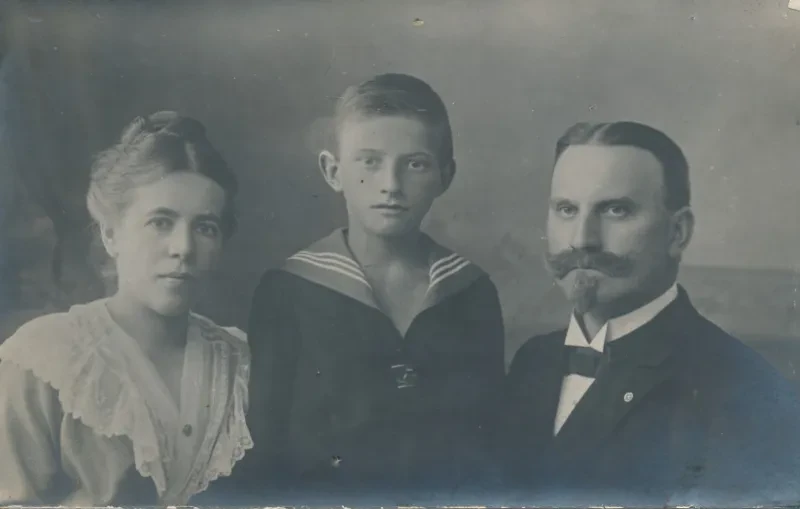 Eine Familie (Mutter, Sohn, Vater) lässt sich fotografieren. Sie tragen die für Anfang des 20. Jahrhunderts typische Kleidung des Bürgertums, der Junge in der Mitte einen Matrosenanzug.